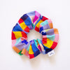 Scrunchie made from fabric with a hand-painted style chequered pattern featuring bright, multicoloured squares in red, yellow, blue, green, pink, and purple, arranged in a circular shape on a white background.