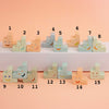 A set of pastel ceramic trinkets numbered 1 through 15, arranged on two white shelves against a peach-pink background. Each block is shaped like a small step stool or chair with a painted smiling face.