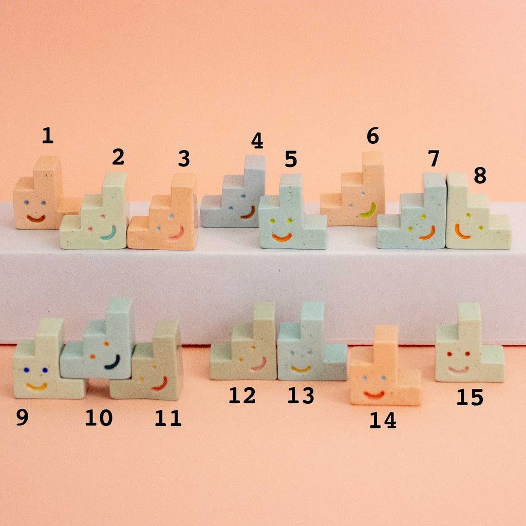 A set of pastel ceramic trinkets numbered 1 through 15, arranged on two white shelves against a peach-pink background. Each block is shaped like a small step stool or chair with a painted smiling face.