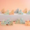 A set of pastel-coloured ceramic trinkets shaped like small steps or stairs, each hand-painted with a simple smiley face. The blocks are arranged in rows on a white platform and against a soft peach-pink background.