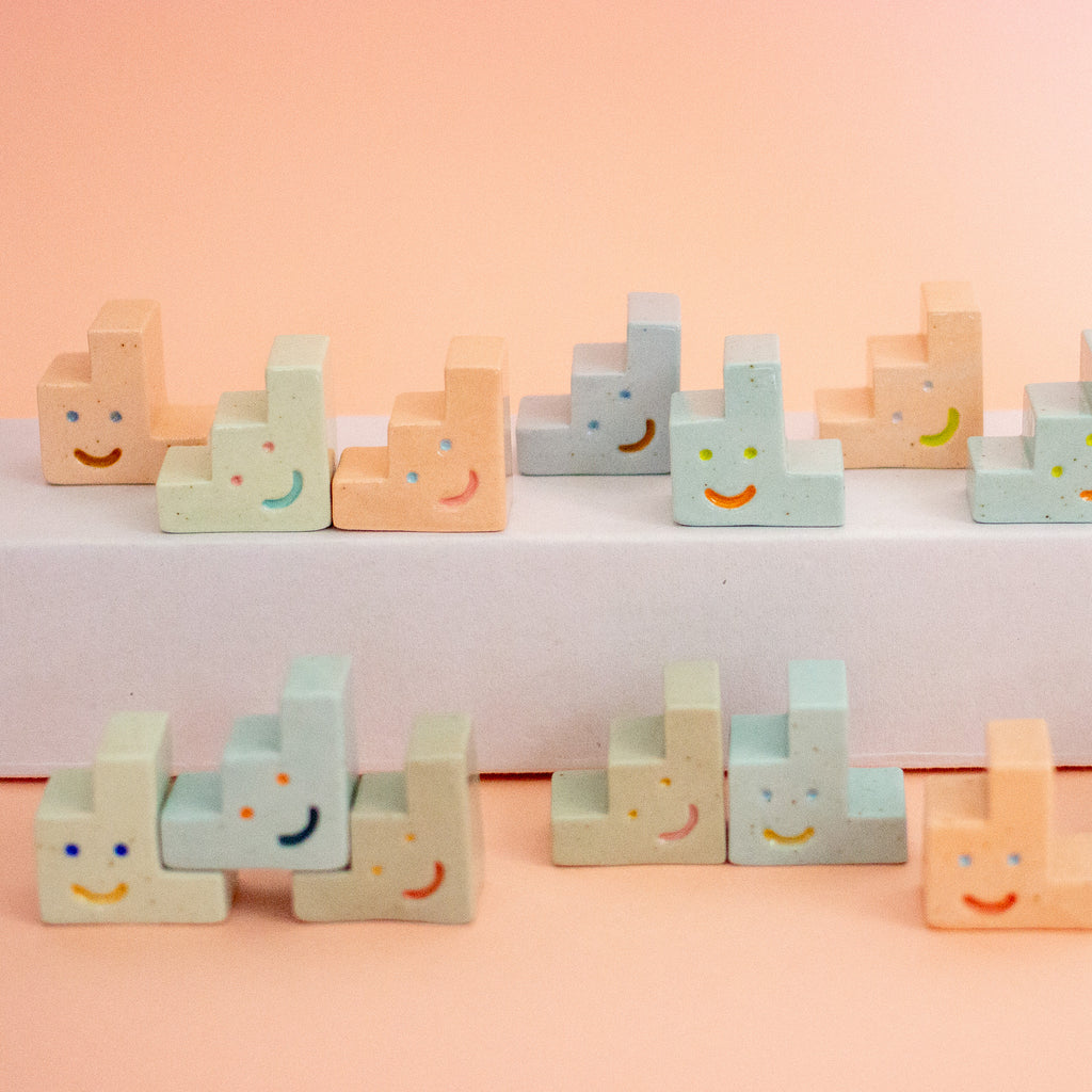 A set of pastel-coloured ceramic trinkets shaped like small steps or stairs, each hand-painted with a simple smiley face. The blocks are arranged in rows on a white platform and against a soft peach-pink background.