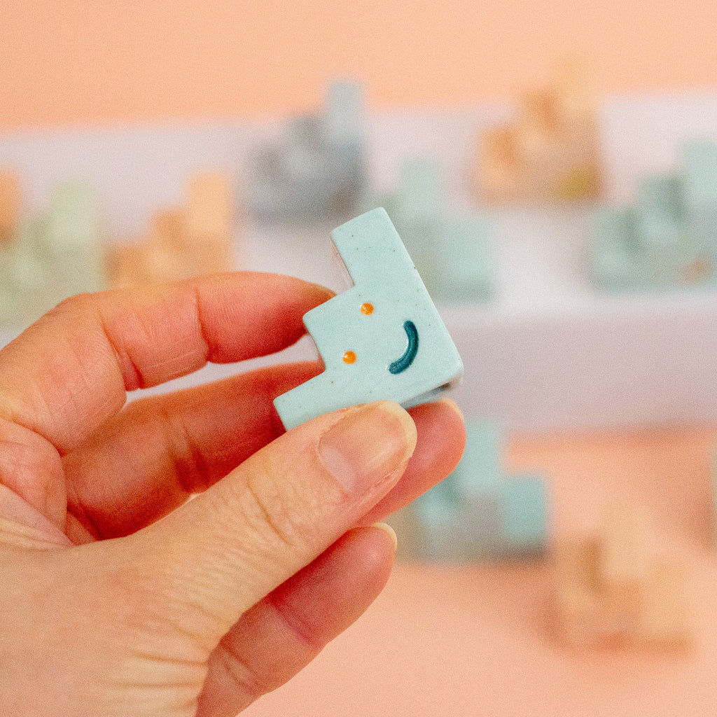 A hand holding a small pastel blue plastic ceramic trinket with a simple smiling face, with more similar blocks blurred in the background against a soft peach-coloured backdrop.