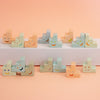 A set of pastel-coloured ceramic trinkets with hand-painted smiling faces, arranged on a white platform against a soft peach-pink background.