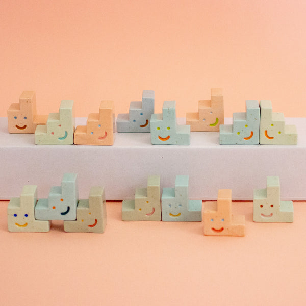 A set of pastel-coloured ceramic trinkets with hand-painted smiling faces, arranged on a white platform against a soft peach-pink background.
