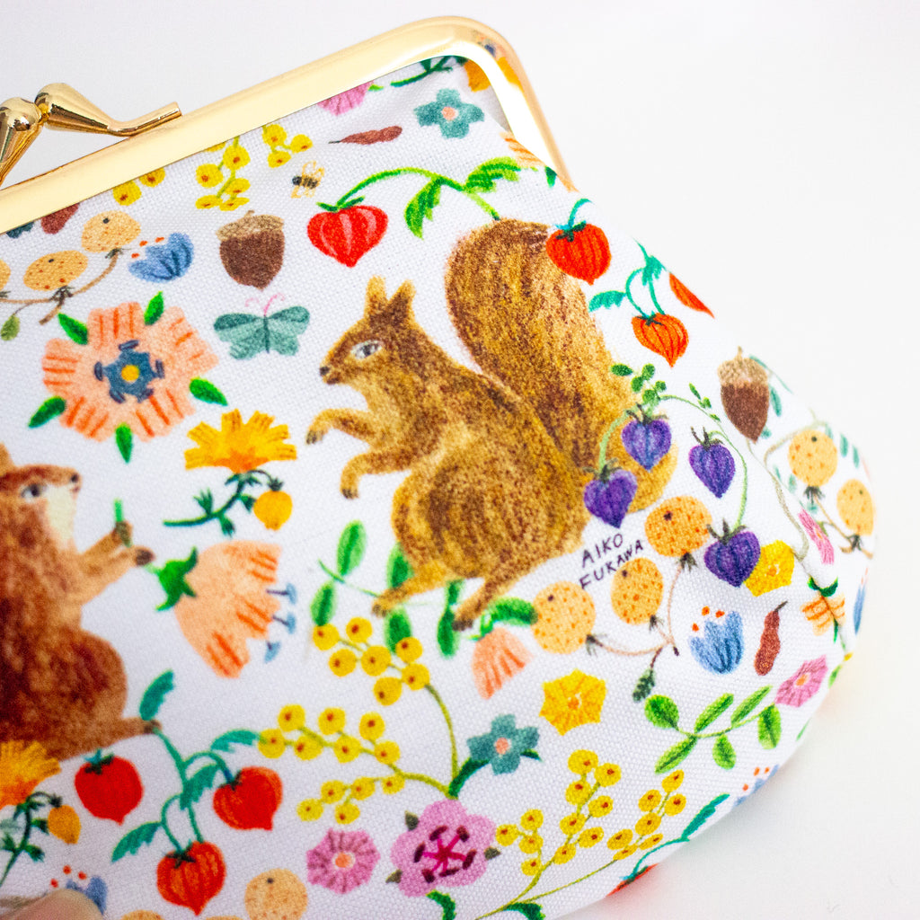 A close-up of a white kiss-lock clutch bag decorated with a colourful print featuring two brown squirrels, red fruits, purple berries, and yellow flowers. The bag has a gold-tone metal frame and is photographed on a clean white background.