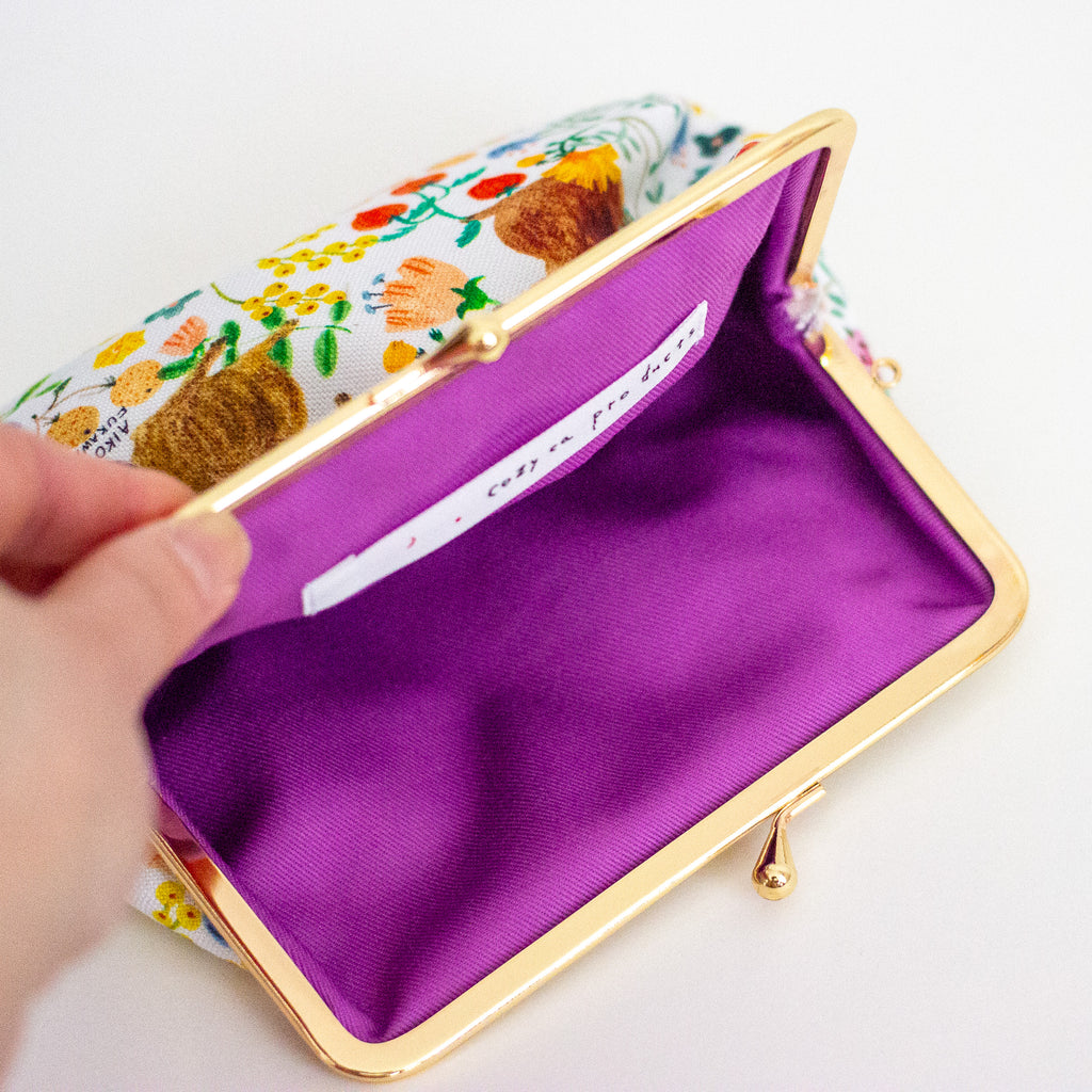 An open kiss-lock clutch purse with a white floral fabric exterior, a gold-tone metal frame, and a bright purple cotton interior. A hand is holding the left side of the purse, and a white interior label is visible.