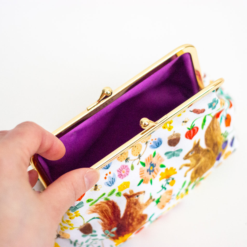 A hand holding an open clutch bag with a white background, colourful floral and squirrel illustrations, a gold-tone metal frame, and a purple cotton interior.