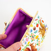 A close-up of a kiss-lock clutch bag with a white exterior decorated with colourful embroidered woodland animals and flowers, shown open to reveal a purple cotton lining. A hand is holding the bag open.