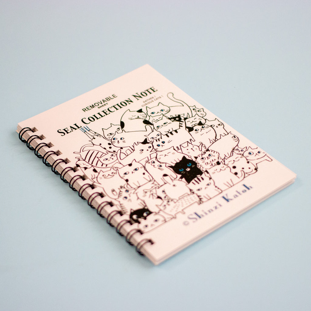 A spiral-bound notebook titled 'SEAL COLLECTION NOTE' with black line-art illustrations of cats on a white cover, photographed on a light blue background.