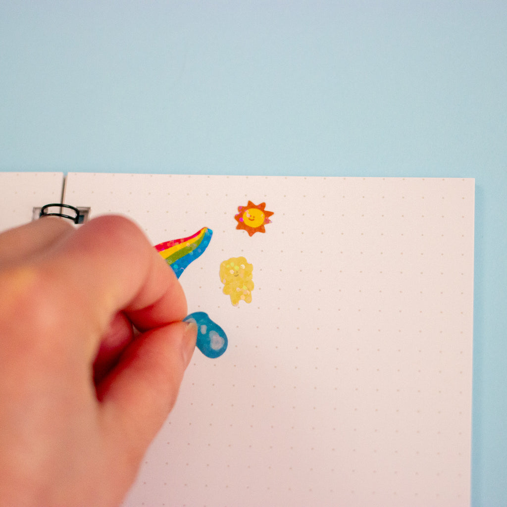 A hand is placing a small blue sticker onto a white dotted page, which is held by a spiral binding against a light blue background. The book already has a rainbow, sun, and yellow flower sticker.