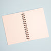 A notebook with white dotted pages and a black spiral coil binding, photographed from above on a light blue background.