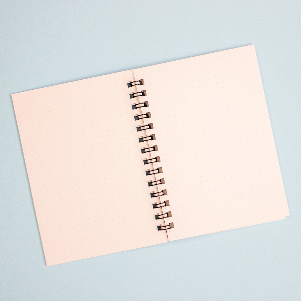 A notebook with white dotted pages and a black spiral coil binding, photographed from above on a light blue background.