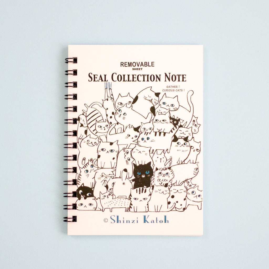 A spiral-bound notebook titled 'SEAL COLLECTION NOTE' with an illustration of many cute cat on the cover, photographed against a pale blue background. The brand name 'Shinzi Katoh' appears at the bottom.