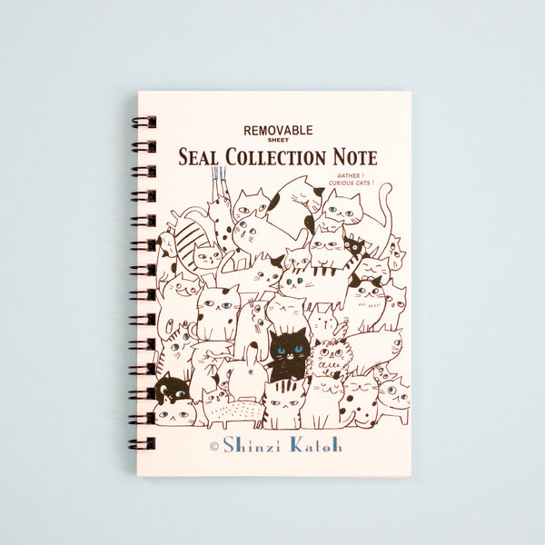 A spiral-bound notebook titled 'SEAL COLLECTION NOTE' with an illustration of many cute cat on the cover, photographed against a pale blue background. The brand name 'Shinzi Katoh' appears at the bottom.