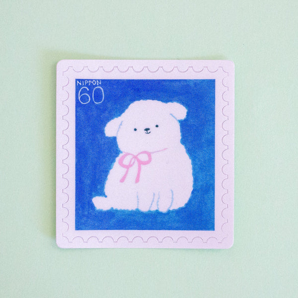 A square postage stamp design featuring a white fluffy puppy wearing a pink bow on a blue background, with a light pink border and the text 'NIPPON 60' in the top left, shown on a pale mint green background.