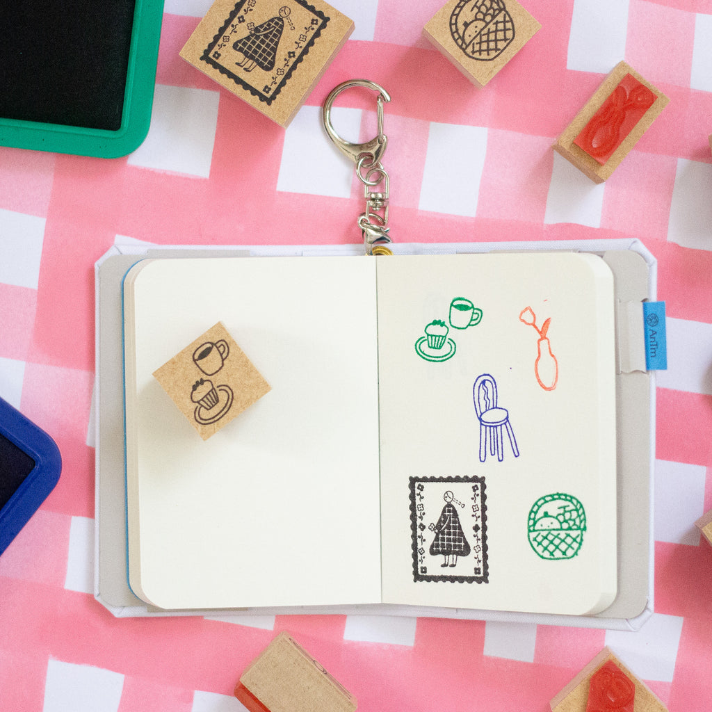 A rubber stamp keychain displayed in an open notebook on a pink and white checkered background, surrounded by other wooden rubber stamps.
