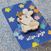 A Misu Juju enamel pin of the bunny character wearing a star on its head and bag, mounted on a blue backing card decorated with colourful stars and the Misu Juju logo.