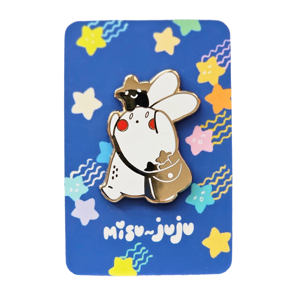 A Misu Juju enamel pin of the bunny character wearing a star on its head and bag, mounted on a blue backing card decorated with colourful stars and the Misu Juju logo.
