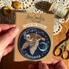 Iron-on patch with owl design held by a hand, surrounded by buttons and thread on a wooden surface.