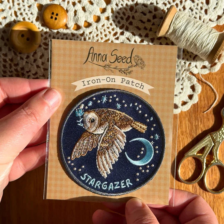 Iron-on patch with owl design held by a hand, surrounded by buttons and thread on a wooden surface.