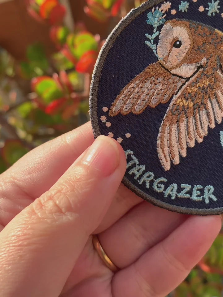 A close-up of a hand holding a round embroidered patch featuring a brown owl with wings spread, on a dark navy background with the word 'STARGAZER' stitched in light blue at the bottom. The background shows blurred outdoor foliage.