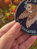 A close-up of a hand holding a round embroidered patch featuring a brown owl with wings spread, on a dark navy background with the word 'STARGAZER' stitched in light blue at the bottom. The background shows blurred outdoor foliage.