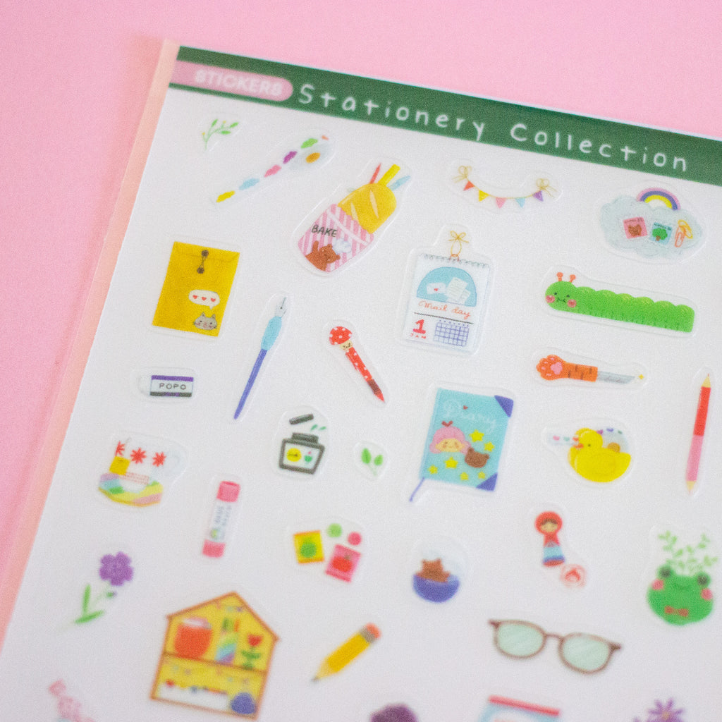 A sheet of colourful stationery stickers displayed on a solid pink background. The sticker sheet contains various cute illustrations of pens, notebooks, flowers, and office items.