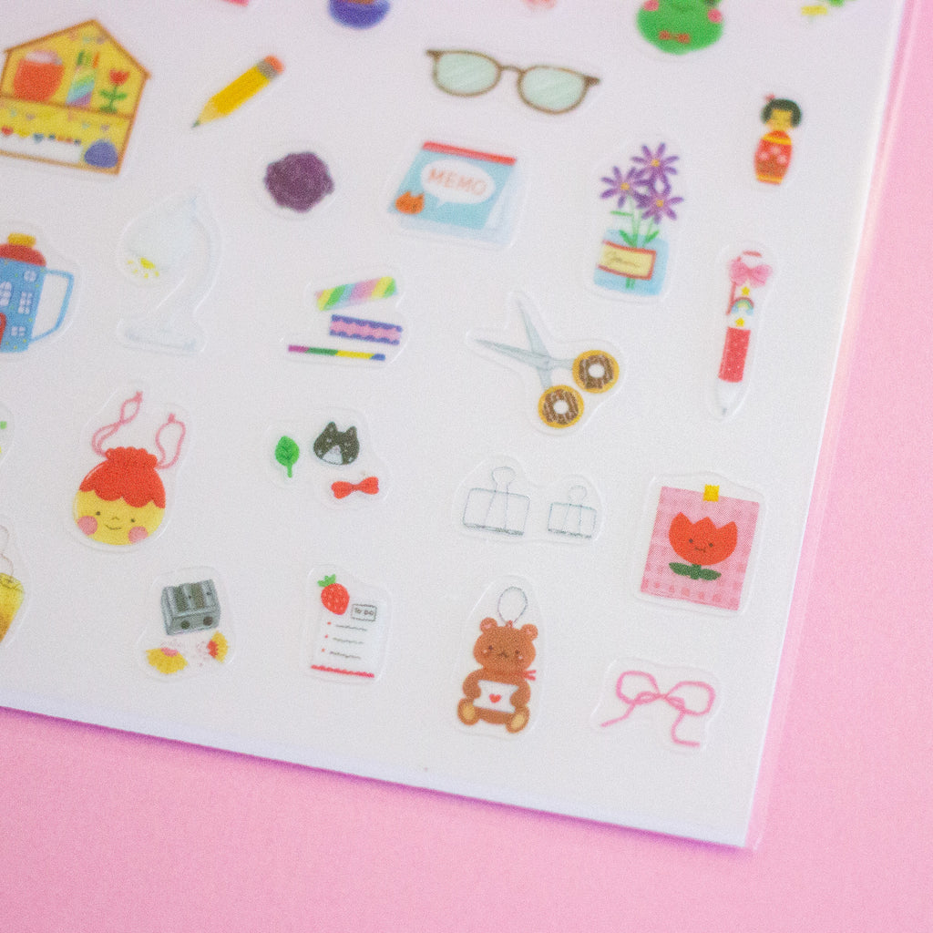 A sheet of colourful stationery stickers displayed on a solid pink background. The sticker sheet contains various cute illustrations of pens, notebooks, flowers, and office items.