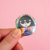 A hand holding a round pinback button badge featuring a stylised cartoon illustration of a woman with black hair and sunglasses, set against a solid pink background.