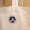 A round pin-back button featuring a cartoon illustration of a person with black hair and sunglasses, set against a colourful striped and checkered background, pinned onto a beige canvas tote bag.