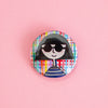 A round pinback button featuring a stylised cartoon illustration of a person with black hair, black sunglasses, and a blue-and-white striped shirt, set against a multicolour plaid background. The button is photographed on a solid pink background.