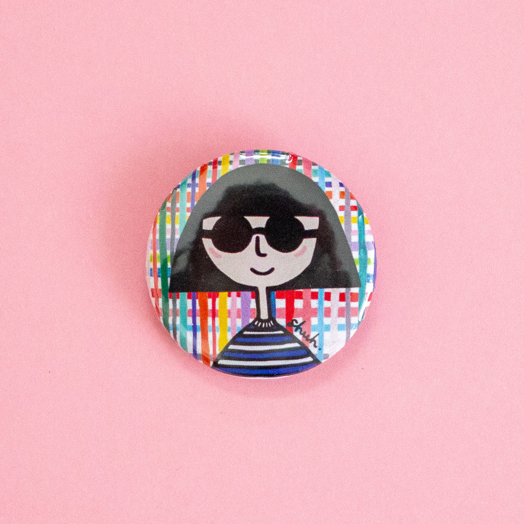 A round pinback button featuring a stylised cartoon illustration of a person with black hair, black sunglasses, and a blue-and-white striped shirt, set against a multicolour plaid background. The button is photographed on a solid pink background.