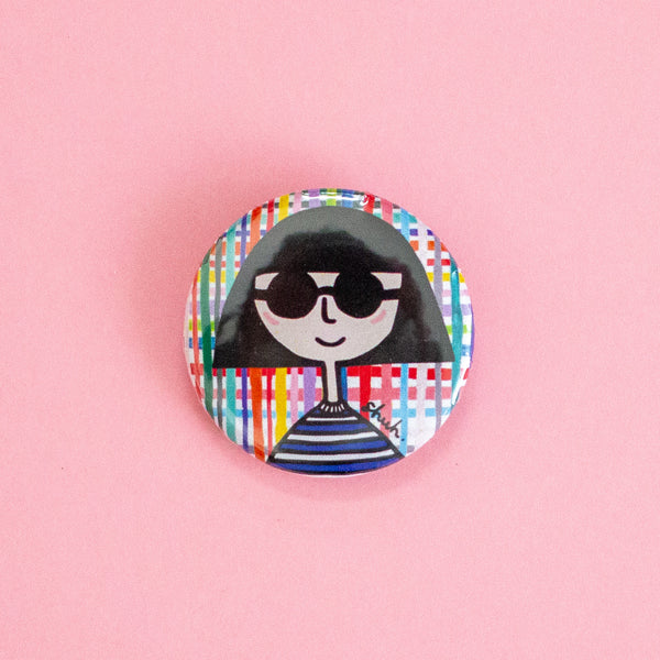 A round pinback button featuring a stylised cartoon illustration of a person with black hair, black sunglasses, and a blue-and-white striped shirt, set against a multicolour plaid background. The button is photographed on a solid pink background.