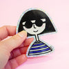A hand holding a die-cut sticker featuring a girl with bob hair wearing sunglasses shaped character with a holographic border, set against a plain pink background.