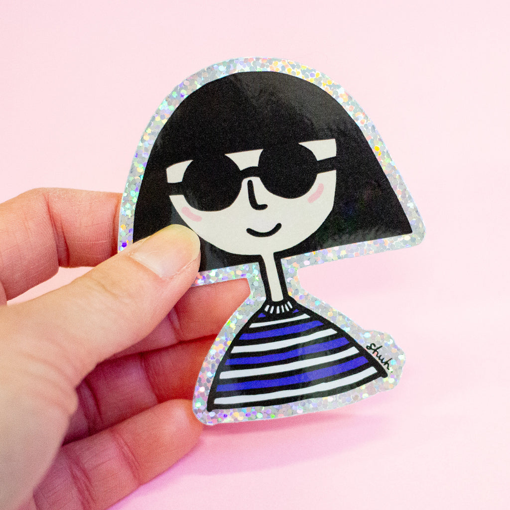 A hand holding a die-cut sticker featuring a girl with bob hair wearing sunglasses shaped character with a holographic border, set against a plain pink background.