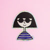 A die-cut sticker featuring a girl with bob hair wearing sunglasses shaped character with a holographic border, set against a plain pink background.