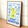 A framed educational poster titled 'STAY HYDRATED' leaning against a white tiled wall on a wooden surface. The poster features a yellow cartoon figure drinking water, blue text, and water droplet illustrations.