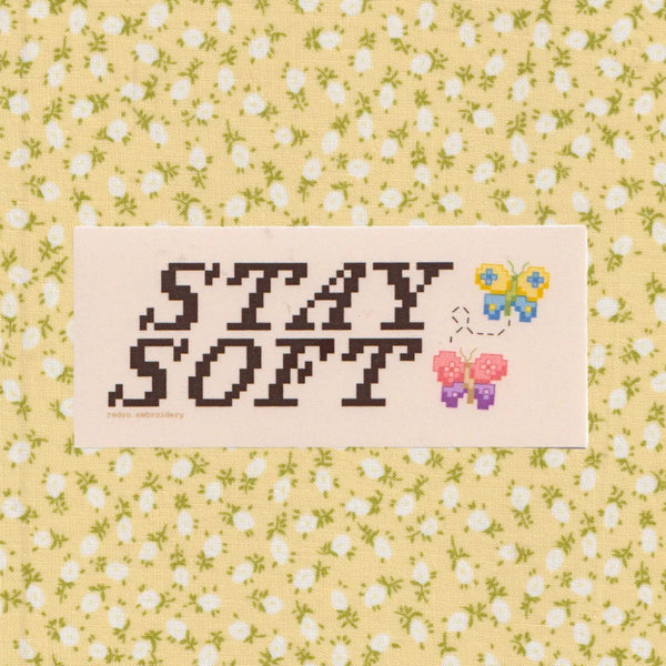 A rectangular cream-coloured patch featuring the words 'STAY SOFT' in dark brown pixel-style lettering, alongside two small pixel-art butterflies in blue/yellow and pink/purple, set against a pale yellow floral background.