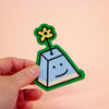 A hand holding a smiling A-shaped character with a green border, light blue body, and a yellow flower on top, set against a solid peach background.