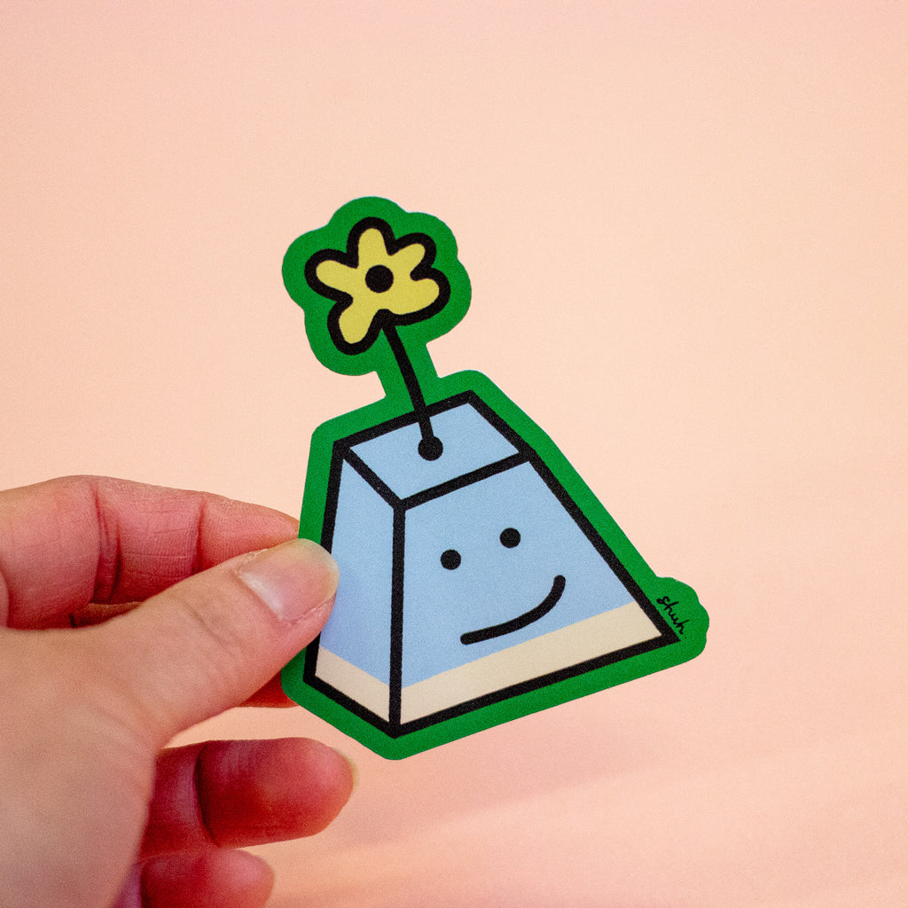 A hand holding a smiling A-shaped character with a green border, light blue body, and a yellow flower on top, set against a solid peach background.