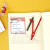 A styled flat-lay of a notebook page with a pink sticky note featuring a cartoon cat and Japanese text, alongside a red compass, a clear ruler, and a pencil on a yellow background.