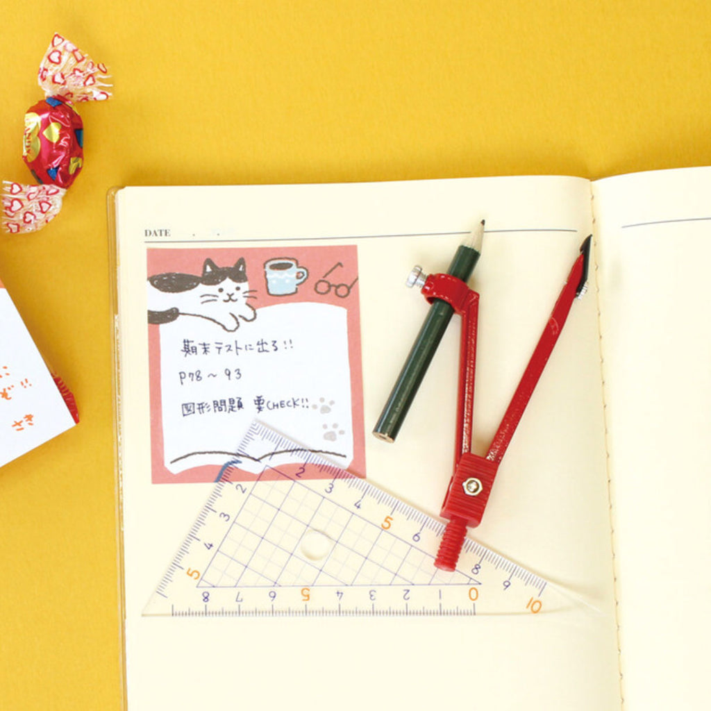 A styled flat-lay of a notebook page with a pink sticky note featuring a cartoon cat and Japanese text, alongside a red compass, a clear ruler, and a pencil on a yellow background.