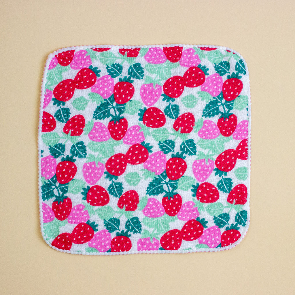 A square fabric item, likely a small towel or cloth, featuring a colourful strawberry pattern on a white background with a white stitched border, set against a solid beige background.