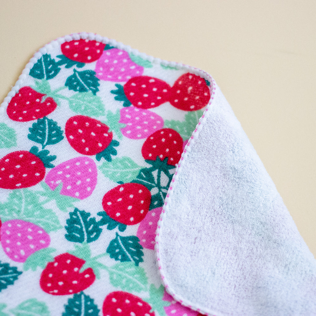 A close-up of a soft fabric towel with a strawberry print on one side and a white plush fleece backing on the other, shown on a plain beige background.