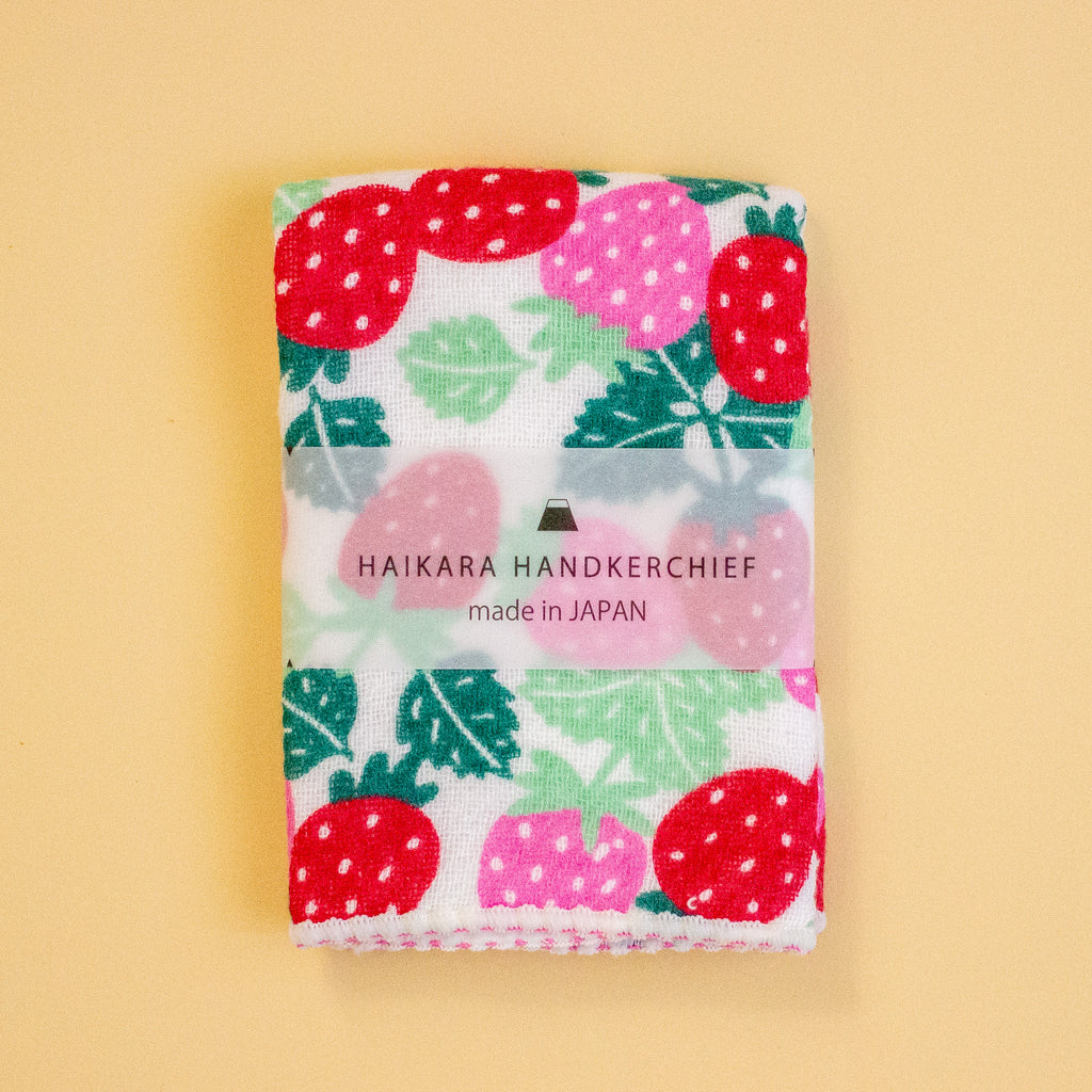 A folded Japanese handkerchief with a strawberry and leaf pattern, featuring a clear paper band label that reads 'HAIKARA HANDKERCHIEF made in JAPAN', set against a solid pale yellow background.