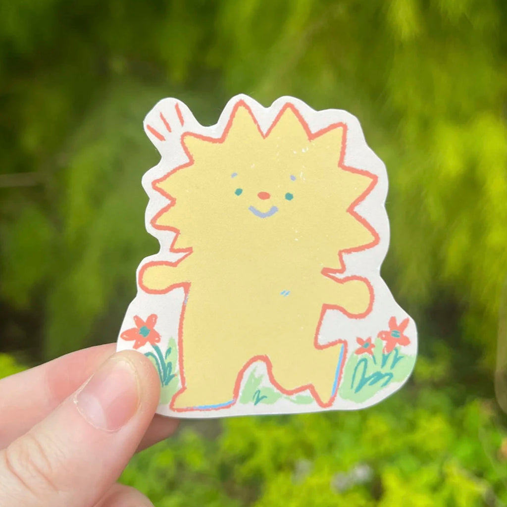 A hand holds a die-cut sticker featuring a cute yellow cartoon creature with red and blue facial details, green grass, and small red flowers, set against a blurred green outdoor background.