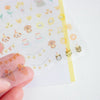 A hand holds a transparent sheet of cute cartoon stickers over a white backing sheet. The stickers feature brown dogs, cats, musical notes, flowers, and birthday cakes in pastel colours.