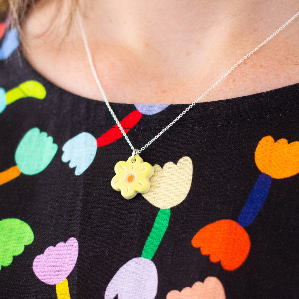 A close-up of a person wearing a delicate silver chain necklace with a small yellow flower pendant, styled over a black top with a colourful tulip pattern.