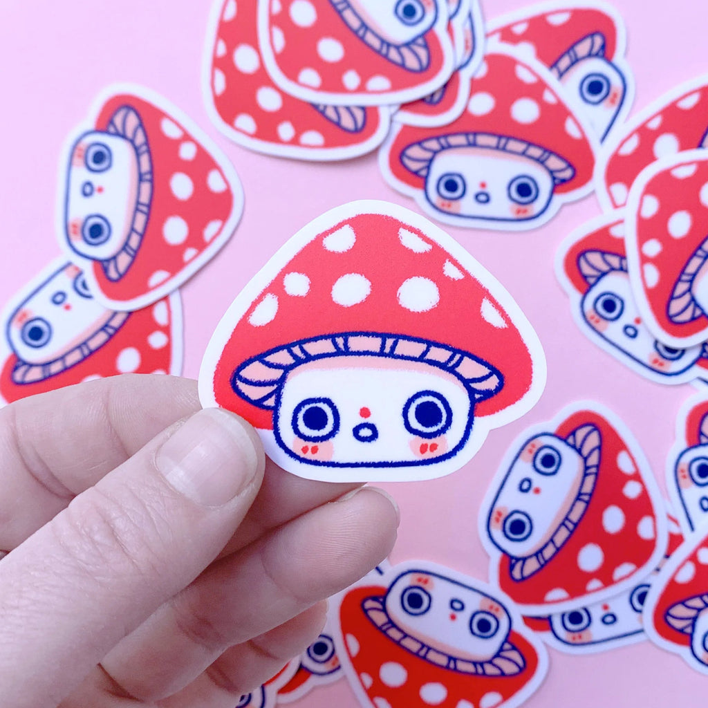 A hand holds a cute red and white polka-dotted mushroom sticker in the foreground, with several identical stickers scattered in the background on a soft pink surface.