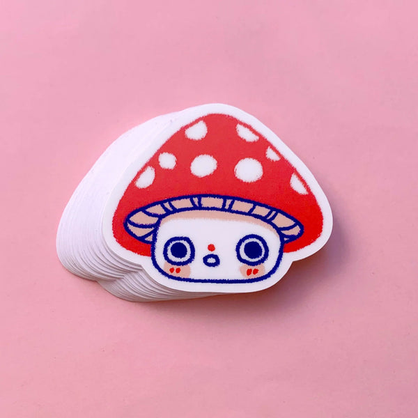 A stack of die-cut stickers featuring a cute cartoon mushroom character with a red cap, white spots, and a white face with blue outlines, displayed on a solid pink background.